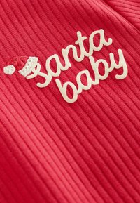 Červená rebrovaná tkanina s vyšitým textom "santa baby" v krémovej farbe, s malou bielou čiapkou Santu nad písmenom "s."