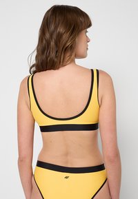 Żółty sportowy biustonosz bikini z czarnymi wykończeniami, z dekoltem w serek na plecach. Materiał wydaje się gładki z minimalnymi detalami metalowymi.