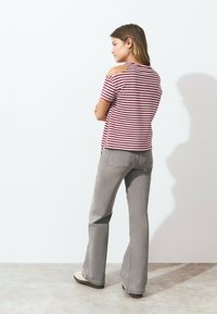 Femme les bras croisés face à un mur blanc, portant un haut à rayures à épaules dénudées, un jean évasé gris et des chaussures blanches.