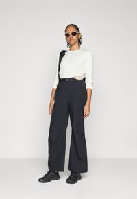 Pantalones cargo anchos negros con bolsillos, combinados con una blusa ligera de manga larga con canalé. Los accesorios incluyen gafas de sol y un bolso de hombro.