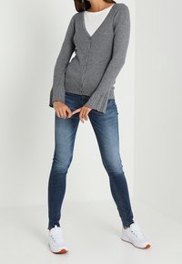 Kvinna iklädd en grå ribbad cardigan med utsvängda ärmar, vit skjorta, blå slim fit-jeans och vita sneakers, stående med benen isär.