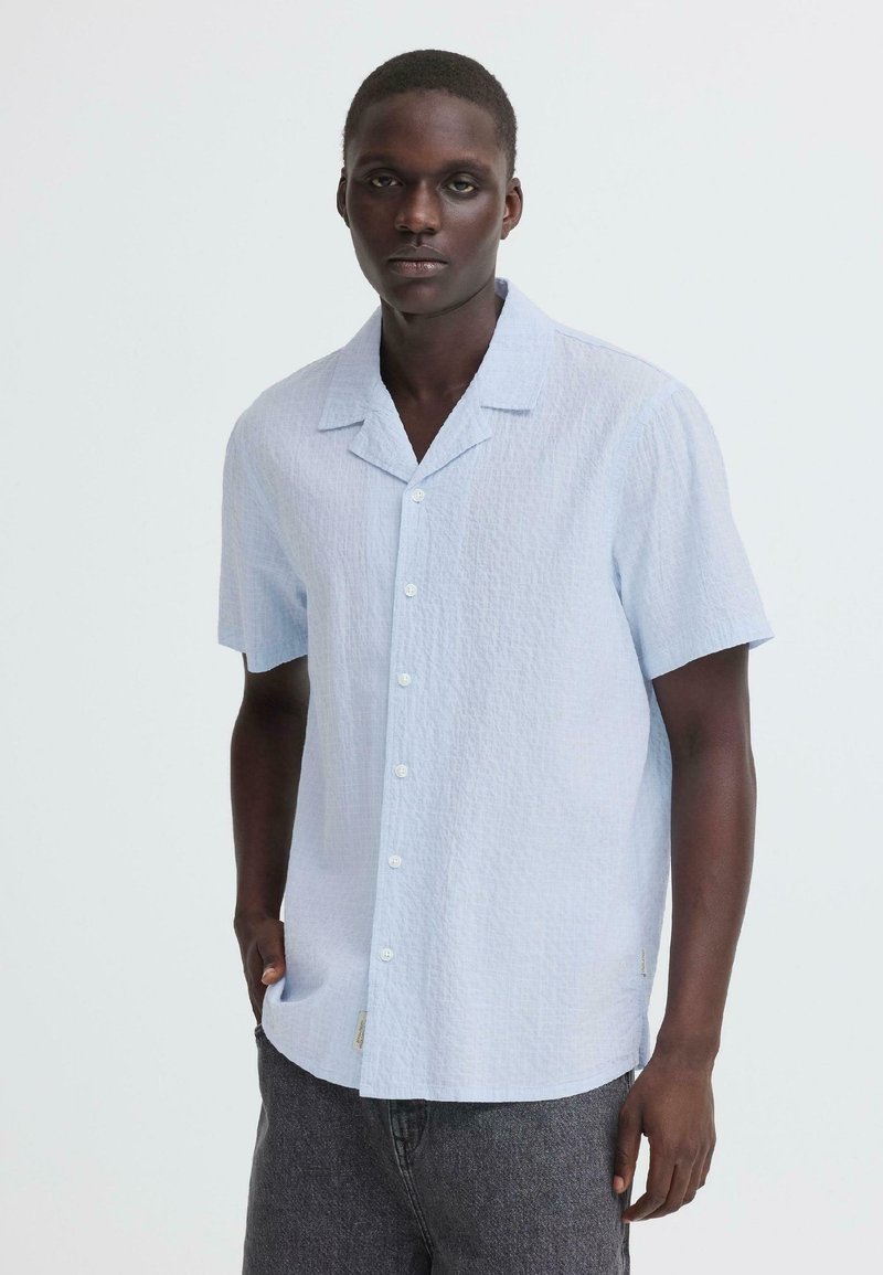 Νεαρός άνδρας που φοράει ανοιχτό γαλάζιο πουκάμισο με κοντά μανίκια και σκούρο γκρι παντελόνι, στέκεται μπροστά σε απλό ανοιχτό φόντο.