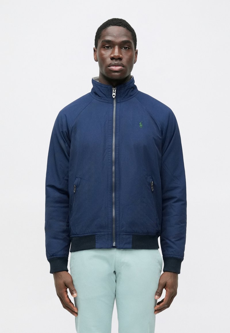 Navy blue zip-up jacket with a high collar, raglan sleeves, and ribbed cuffs; features a green logo on the left chest and two side pockets.