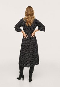 Černé maxi šaty s puntíky, s úzkým pasem, tříčtvrtečními zvonovými rukávy a hladkou texturou, doplněné černými kozačkami ke kolenům.