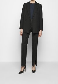 Svart dubbelknäppt kavaj med en strukturerad passform, i kombination med skräddarsydda svarta byxor med sidofickor och spetsiga svarta högklackade skor.