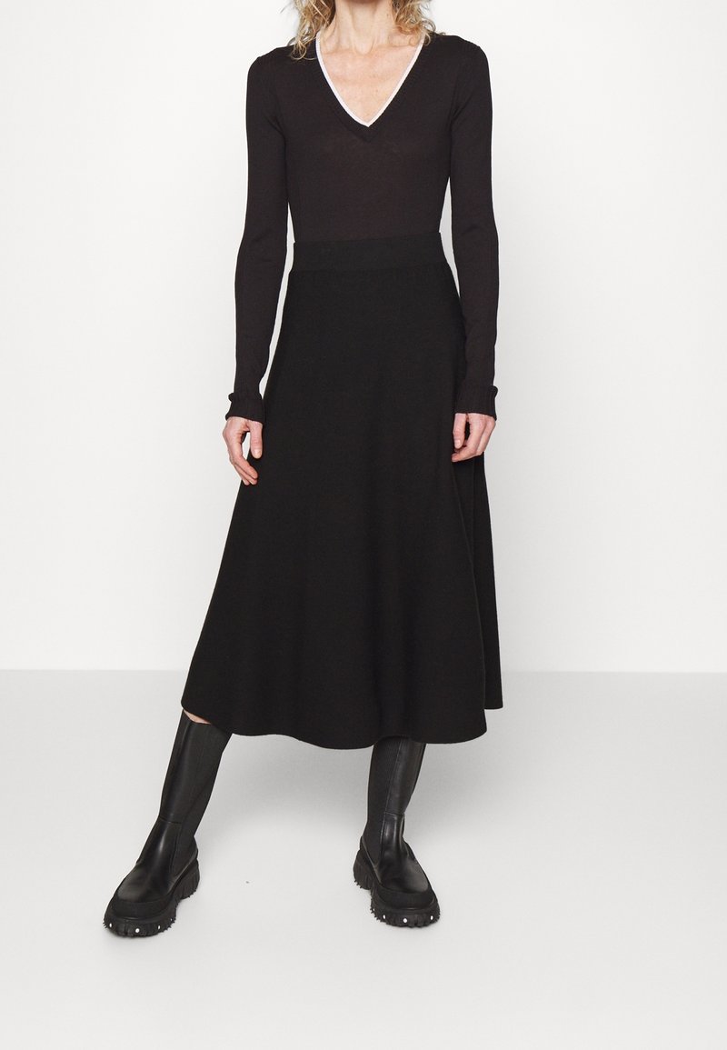 Zwarte top met lange mouwen en een V-hals, met contrasterende randen, gecombineerd met een zwarte lange rok, gedragen met zwarte kniehoge laarzen.