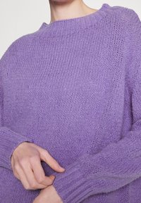 Personne portant un pull en tricot violet, ajustant le poignet d'une manche avec la main opposée.