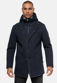 Mørkeblå frakke med en struktureret design, der har en hætte, kontraststing, fem knapper og to forlommer. Tekstureret stof er synligt.