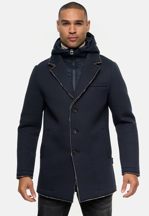 Cappotto blu navy con un design strutturato, dotato di cappuccio, cuciture a contrasto, cinque bottoni e due tasche frontali. Tessuto testurizzato visibile.