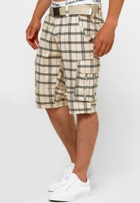Bež karirane cargo kratke hlače s crnim mrežastim linijama, s bočnim džepovima i kontrastnim bijelim pojasom Calvin Klein. Nosene s bijelim tenisicama.