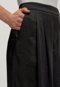 Zwarte geplooide broek met een gladde textuur, voorzien van zijzakken en een hoge tailleband. De stof lijkt lichtgewicht en mat.