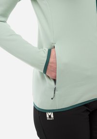 Veste en polaire vert clair avec une poche zippée, bordure contrastante vert foncé et texture lisse, associée à un pantalon noir avec un logo.