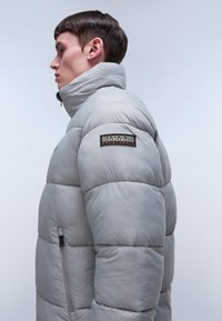 Grå puffjacka med hög krage, med kviltade sektioner, en ficka med dragkedja och en logotyp på vänster axel.