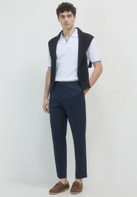 Homme portant un polo blanc à manches courtes, un pantalon bleu marine, des mocassins marron et un pulls noir drapé sur les épaules, se tenant contre un fond blanc.
