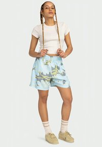 Jovem com tranças longas segura as pontas do cabelo, vestindo uma t-shirt branca curta, calções azuis com padrão floral, sapatos bege e meias às riscas.