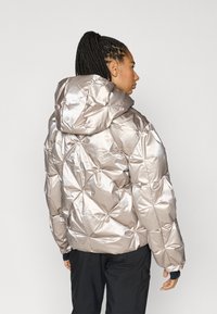 Spīga sudraba jaka ar metālisku spīdumu, ar kapuci, ģeometrisku šuvumu un elastīgiem aprocēm, valkāta pār melniem apģērbiem.