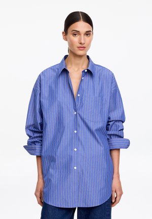 Žena s tmavými vlasmi zviazanými dozadu nosí voľnú modrú košeľu s dlhým rukávom a bielymi a oranžovými zvislými pruhmi.