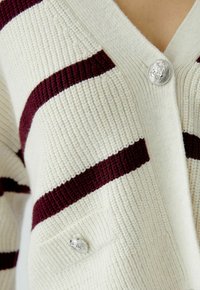 Cremfarbener und burgunderfarbener gestreifter Cardigan mit gerippter Textur, ausgestattet mit silbernen Löwenknöpfen und einem V-Ausschnitt.
