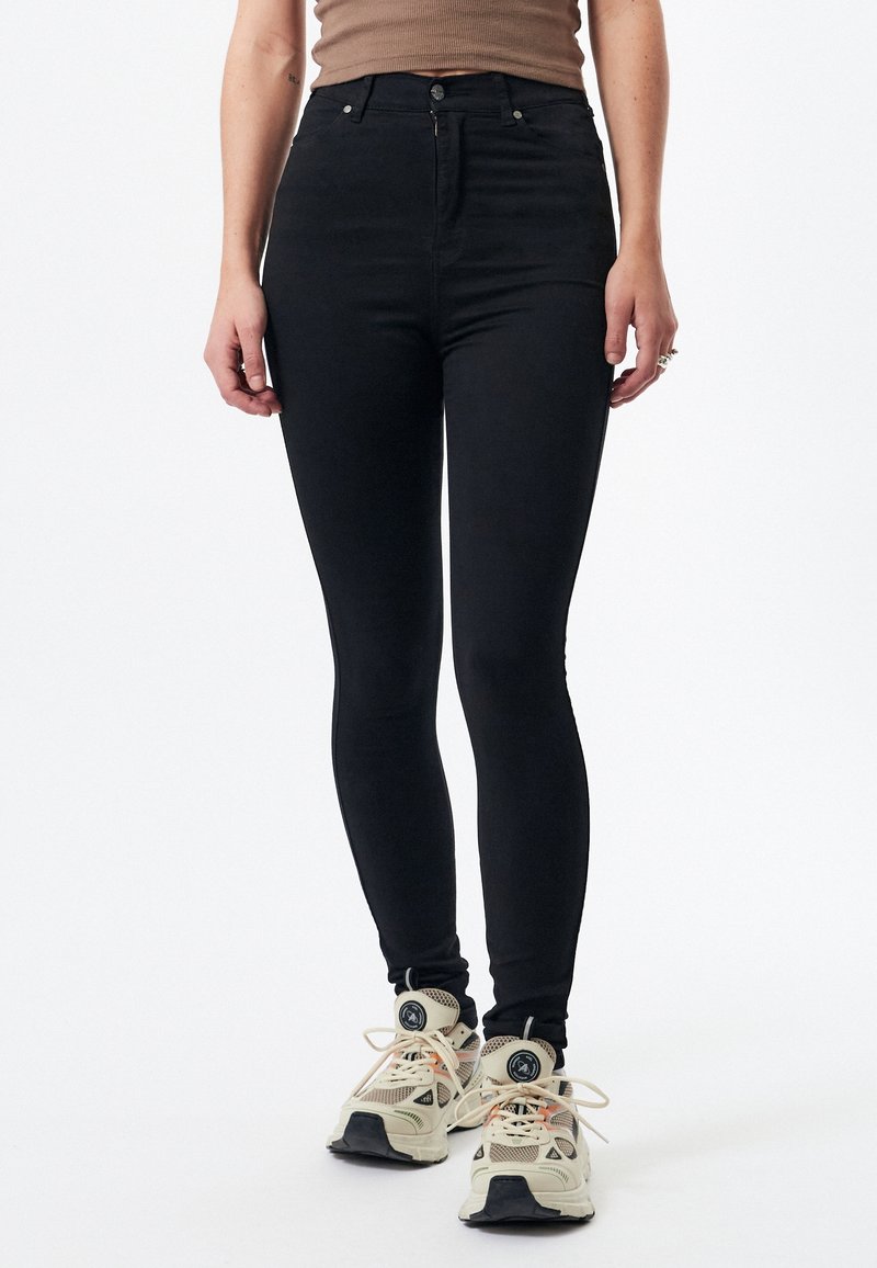 Jean noir taille haute avec une coupe slim, comportant cinq poches, des passants de ceinture et une fermeture à boutons. Portés avec des baskets de couleur claire.