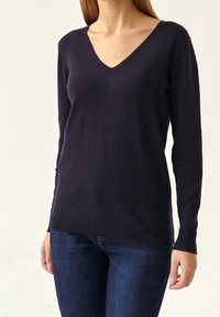 Marineblaues langärmliges V-Ausschnitt-Pullover aus weichem Stoff mit gerippten Bündchen und Saum. Über dunklen Jeans getragen, was eine figurschmeichelnde Silhouette zeigt.