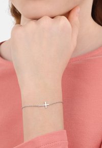 Bracciale in argento con un semplice ciondolo a croce, indossato su una mano con una manica rosa pallido sullo sfondo. Finitura metallica liscia.