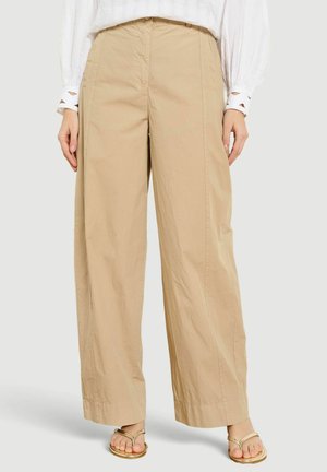 Beżowe spodnie z szerokimi nogawkami noszone z białą bluzką z długim rękawem i złotymi sandałami na otwarte palce, pokazane od talii do stóp.