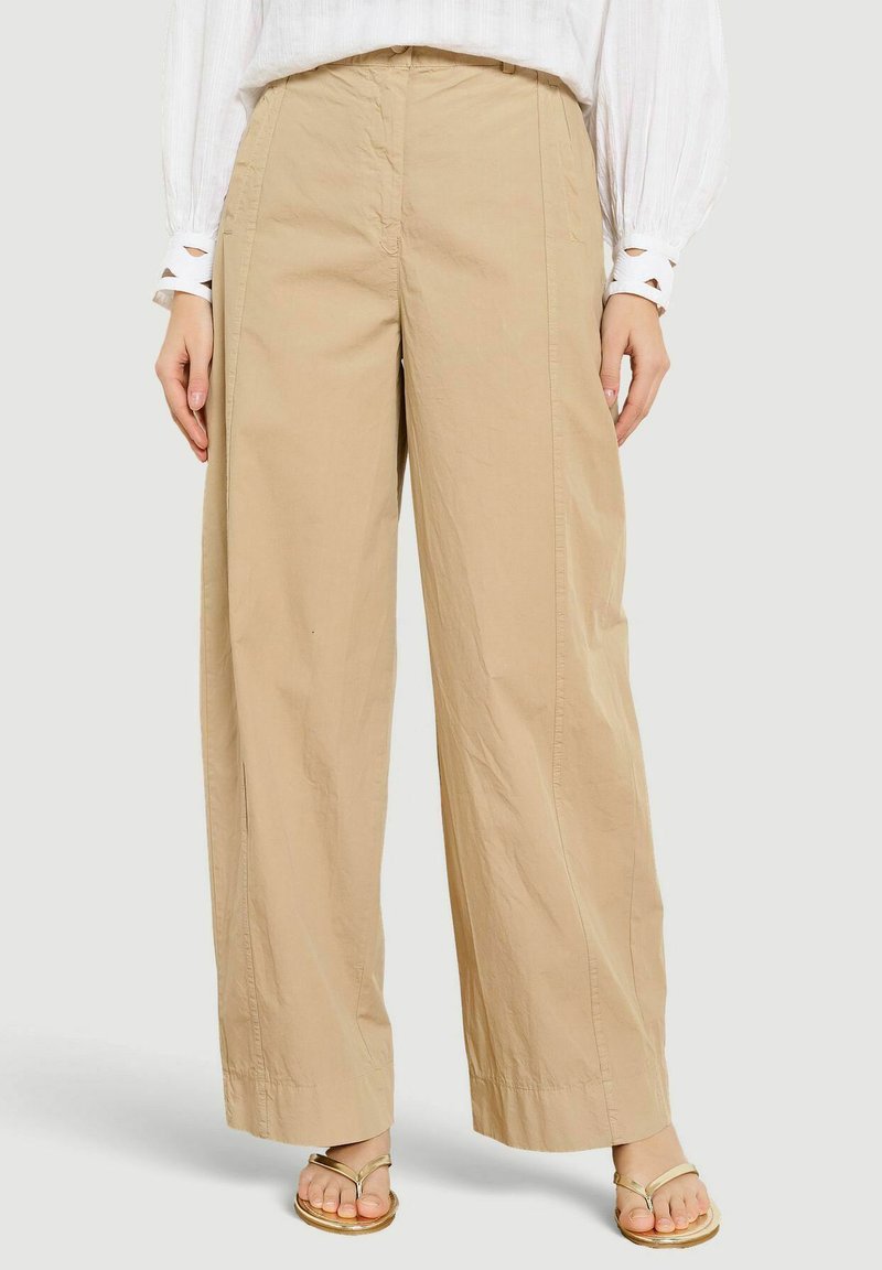 Béžové nohavice s širokými nohavicami nosené s bielym tričkom s dlhými rukávmi a zlatými sandálmi s otvorenou špičkou, zobrazené od pása po chodidlá.