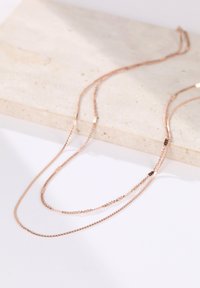 Tres delicadas cadenas de oro rosa de diferentes longitudes, con eslabones suaves y pequeños acentos geométricos, descansando sobre una superficie de piedra texturizada.