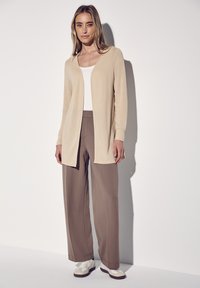 Cardigan lungo beige realizzato in tessuto morbido, indossato sopra un top bianco e pantaloni brown larghi. L'outfit è abbinato a sneaker bianche.