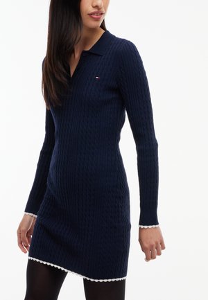Frau trägt ein marineblaues, langärmeliges Strickkleid mit weißer, geschwungener Einfassung und schwarzen Strumpfhosen vor weißem Hintergrund.