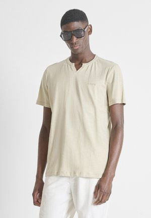 Giovane uomo che indossa una camicia beige a maniche corte e pantaloni bianchi, in piedi davanti a uno sfondo chiaro e uniforme, con grandi occhiali da sole neri.