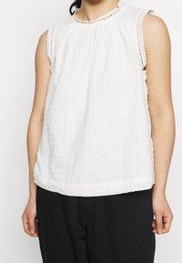 Vit ärmlös topp med en veckad design, texturerad med små pom-pom-mönster, och en spetsprydd halsringning. Matchad med mörka byxor.