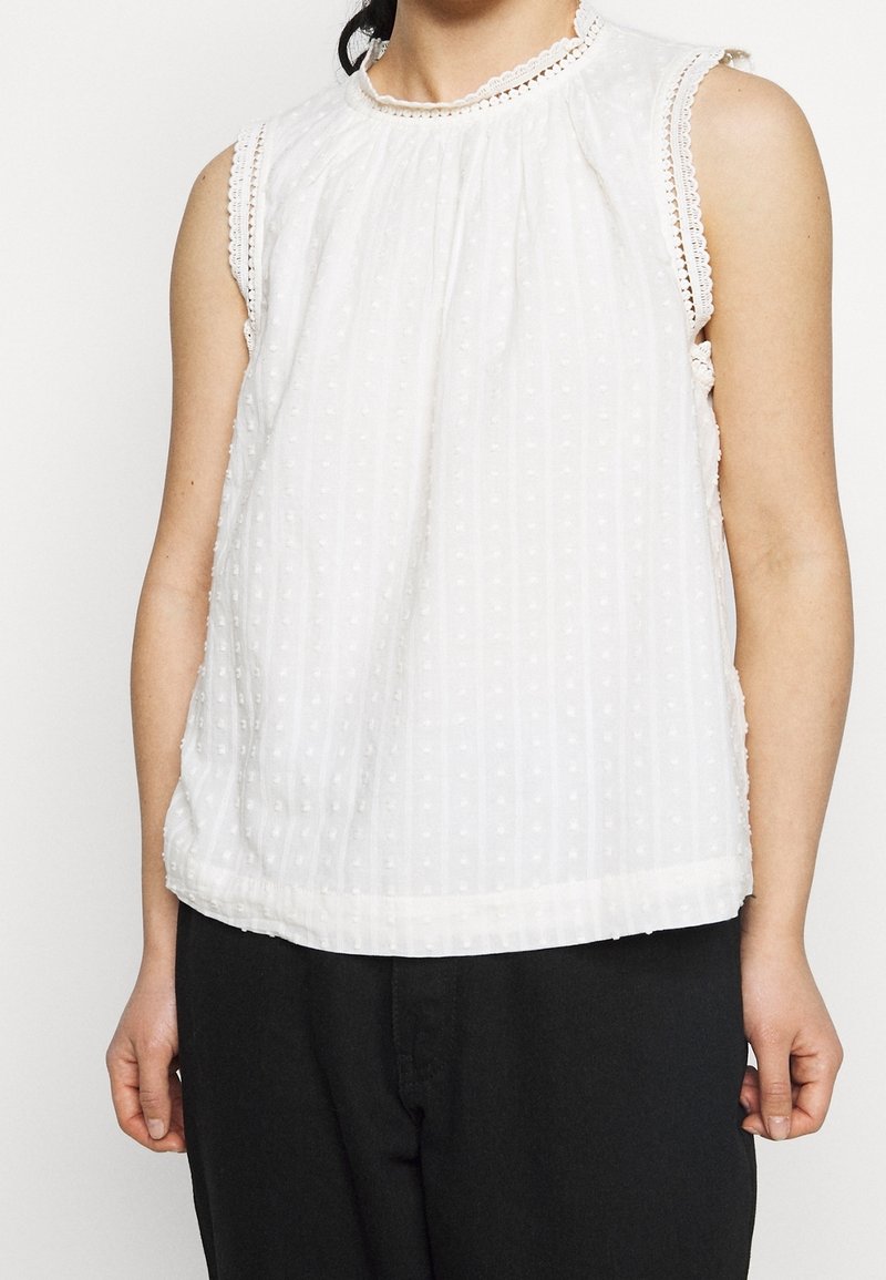 Vit ärmlös topp med en veckad design, texturerad med små pom-pom-mönster, och en spetsprydd halsringning. Matchad med mörka byxor.