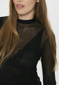 Top negro de manga larga hecho de tela transparente con un patrón texturizado. Presenta un cuello alto y un look en capas sobre un sujetador negro.