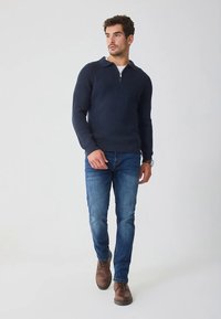 Σκούρο μπλε πλεκτό πουλόβερ με φερμουάρ και γιακά. Φοράει μπλε τζιν και καφέ δερμάτινα παπούτσια με κορδόνια, μοντέλο στέκεται σε ανοιχτό φόντο.