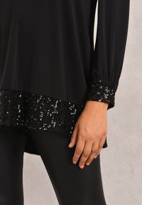 Black long-sleeve top with a sequin-embellished hem and cuffs; smooth fabric; relaxed fit, with textured black leggings below.