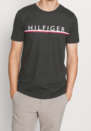 Homme portant une chemise à manches courtes gris foncé avec le texte "HILFIGER" et des rayures rouges et blanches au niveau de la poitrine, associée à un pantalon beige.