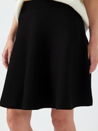 Femme portant une jupe noire ample longueur genou et un haut blanc, se tenant les mains détendues sur les côtés devant un fond uni.