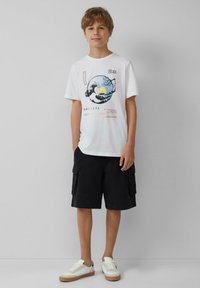 Weißes Baumwoll-T-Shirt mit einem bedruckten Wellen- und Sonnenaufgangsdesign, kombiniert mit schwarzen Cargo-Shorts und weißen Sneakers mit beigen Akzenten.