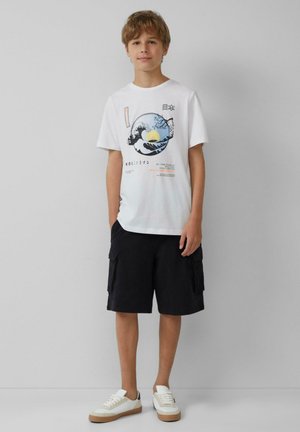 Witte katoenen t-shirt met een bedrukte golf- en zonsopgangontwerp, gecombineerd met zwarte cargoshorts en witte sneakers met beige accenten.