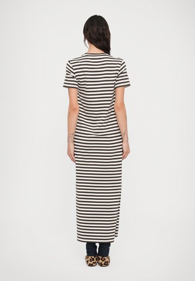 Γυναίκα που στέκεται με την πλάτη προς την κάμερα, φοράει μακρύ φόρεμα με καφέ και λευκές ρίγες, σκούρο παντελόνι και παντόφλες με σχέδιο λεοπάρ.