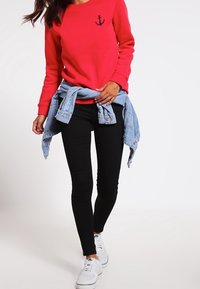 Červená mikina s potiskem kotvy, černé legíny a džínová bunda uvázaná v pase. Celý outfit doplňují bílé tenisky.