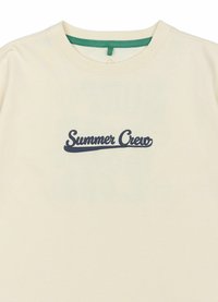 Camiseta de algodón color crema con un logo en cursiva "Summer Crew" en color azul marino centrado en el pecho y un acento verde en el cuello.