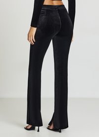 Pantalon noir en velours évasé avec fentes latérales, texture lisse et taille ajustée. L'arrière présente des détails de coutures discrètes et une coupe classique.
