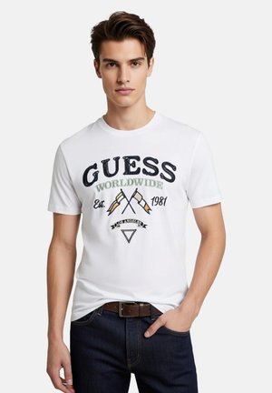 Jeune homme portant un T-shirt blanc Guess avec des inscriptions en noir et vert, des drapeaux croisés et le texte "Los Angeles", assorti d'un jean foncé et d'une ceinture marron.