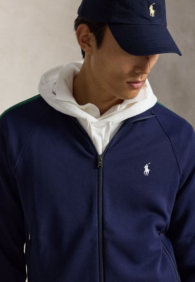 Homme portant une veste zippée bleu marine et une casquette assortie avec un logo blanc, superposée à un sweat à capuche blanc, regardant vers le bas et sur le côté.
