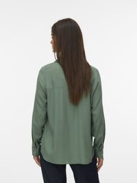 Chemise à manches longues en tissu vert forêt, avec un empiècement au dos, des poignets à boutons et une texture lisse. Portée avec un jean foncé.