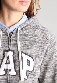 Felpa grigia con zip, dotata di lacci bianchi e grande logo "GAP" ricamato. Presenta una texture morbida e melange e un cappuccio.