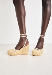 Espadrilles de cuña con un exterior tejido en color beige y correas ajustables blancas con tachuelas doradas. Superficie texturizada y diseño de punta redonda.