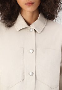 Woman wearing a beige button-up jacket with two chest pockets and curly dark hair visible near the collar.